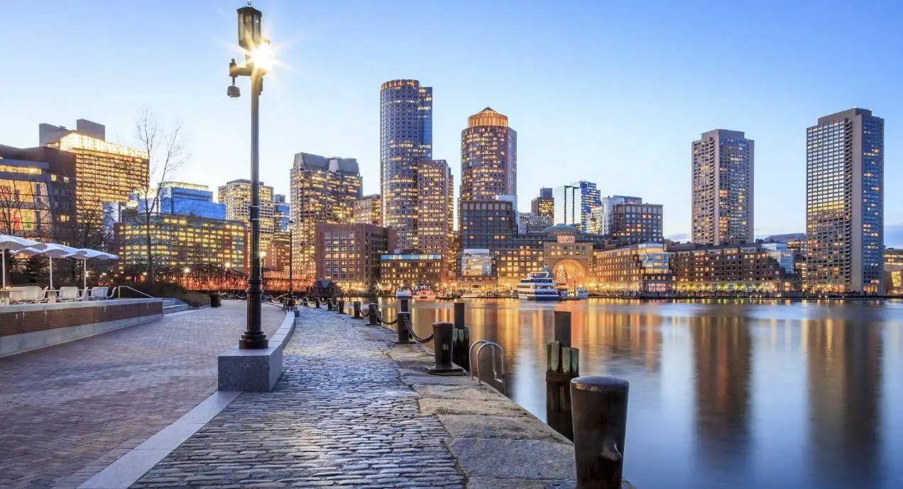 Boston's Harborwalk at dusk, with a cobblestone waterfront path and lamppost leading toward the illuminated downtown skyline across the harbor.