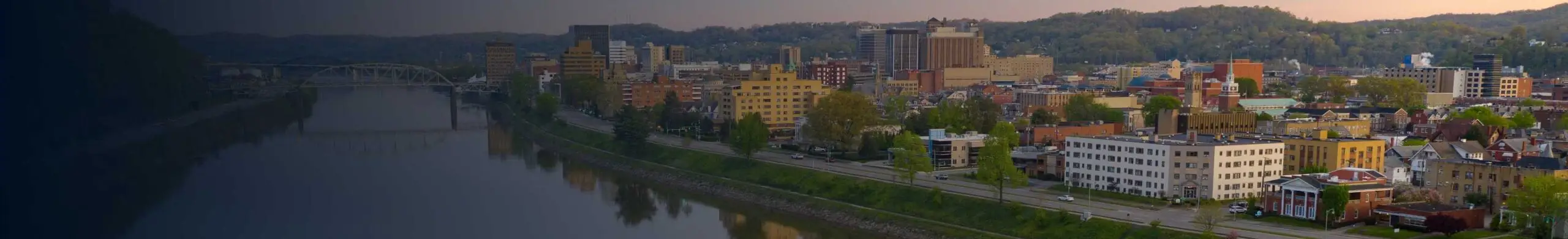 Aerial panoramic view of a small Appalachian city skyline along a curving river, with a truss bridge, downtown buildings, and tree-covered hills in the background at dusk.