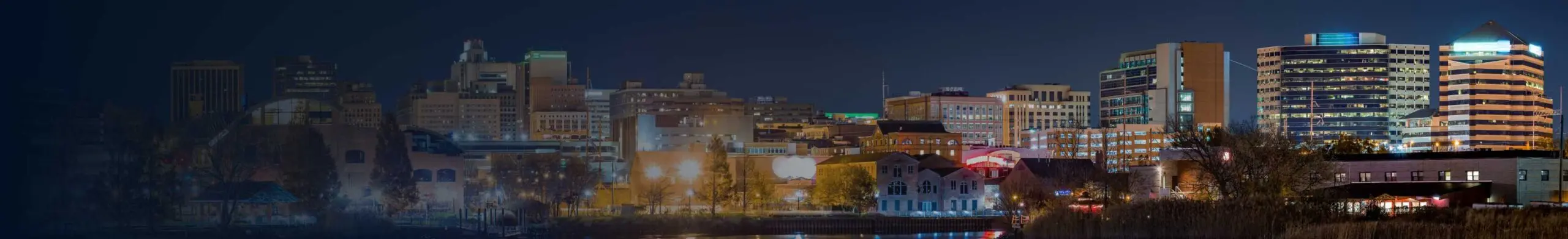 Panoramic night view of a mid-sized city skyline lit up along a riverfront, with a mix of historic brick buildings and modern high-rises against a dark night sky.