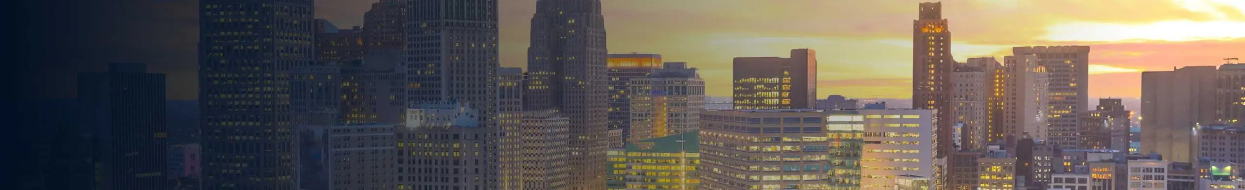 Panoramic aerial view of a dense downtown skyline at sunset, with a mix of historic and modern high-rise buildings lit up against a glowing orange sky.