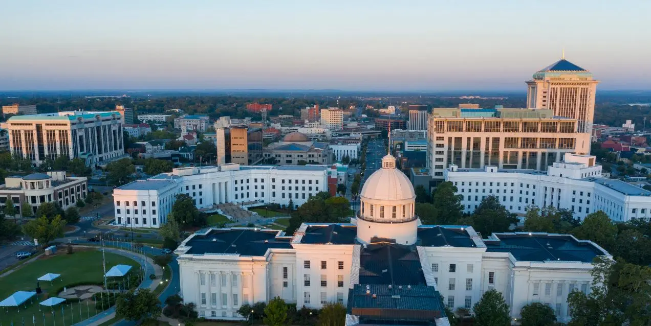 Aerial view of Montgomery, Alabama, showing the Alabama State Capitol's domed building in the foreground with the downtown skyline stretching behind it at dusk.
