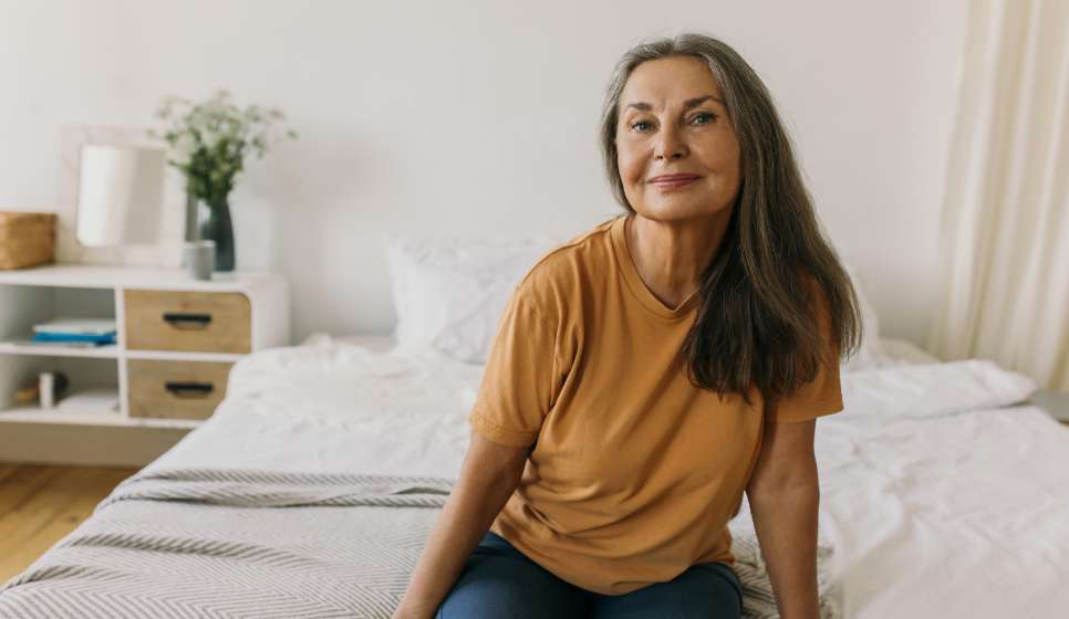 A middle-aged woman sitting comfortably in a bright bedroom.