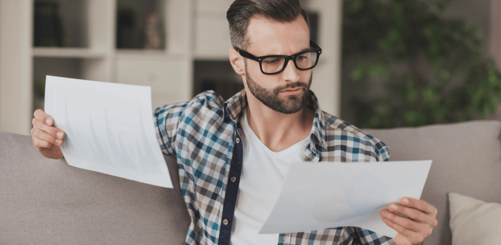 Man holding two pieces of paper and looking at one of them while sitting on a beige couch